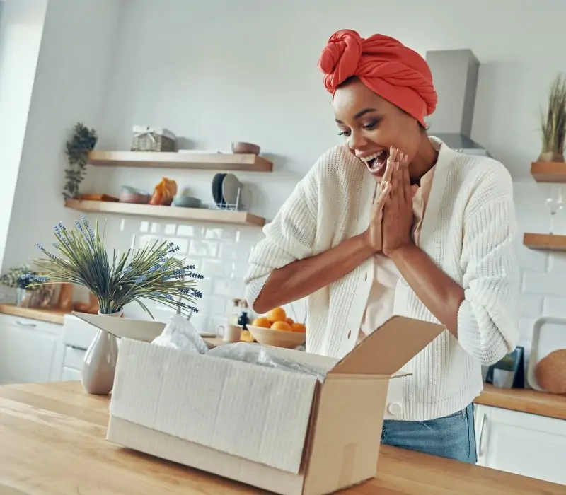 Excited African woman looking at the opened box and clapping hands while standing at kitchen